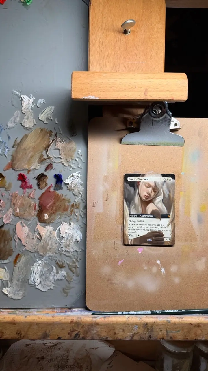 A wooden tabletop easel holds a small trading card on a clip. The card is being modified with painted artwork showing soft, light-toned wings and a partially rendered figure. To the left of the easel is a painter’s palette covered in mixed oil paints—mostly neutral tones like beige, browns, whites, and touches of bright colors such as red and blue. A thin paintbrush rests horizontally along the bottom edge of the setup.