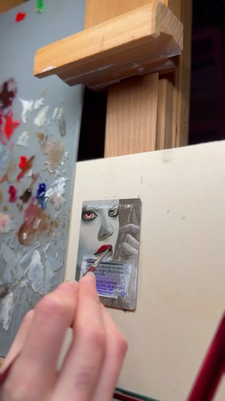 A hand holds a fine paintbrush and paints on a trading card attached to a wooden easel. The artwork on the card depicts a stylized face with pale skin, vivid red lips, and intense red eyes. The artist is applying detail to the lips. To the left is a palette covered in mixed oil paints in various neutral and vivid hues.
