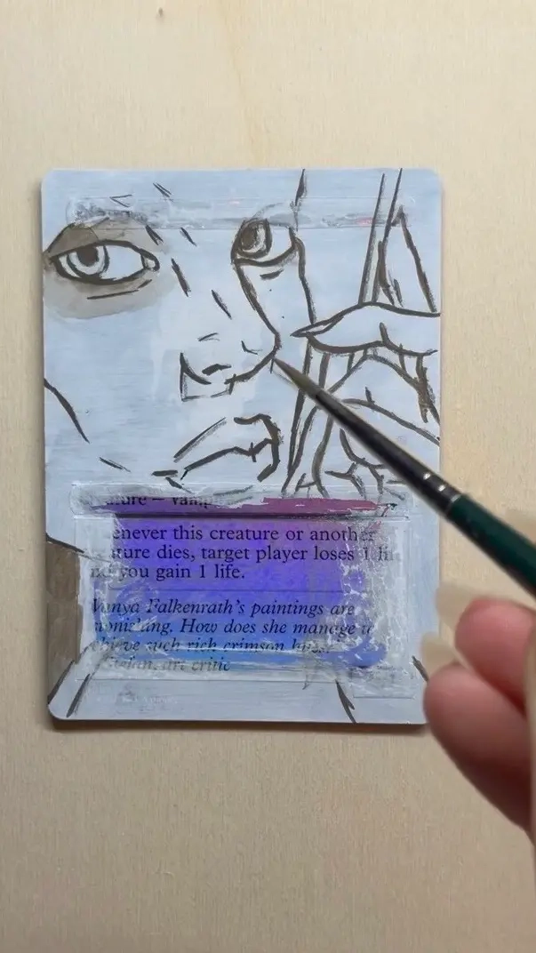 A trading card lies on a flat surface while someone paints over it with a small brush. The card has a rough sketch of a face and hand drawn in dark lines over a lightly colored background, suggesting the early stages of a character illustration. The text area of the card remains partially visible beneath a layer of paint.